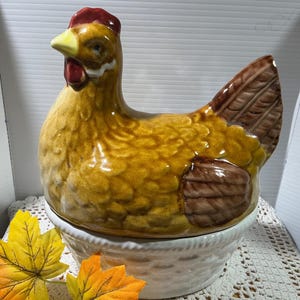 Op de afbeelding: Een keramische kipvormige container met een bruin en geel glazuur. De kip heeft een rode kam en lellen en staat op een witte mandweefbasis. Decoratieve herfstbladeren zijn bij de basis geplaatst.