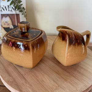Peut inclure: Un sucrier vintage avec couvercle et un pot à crème. Les deux pièces ont un glaçage bicolore marron et beige. Le sucrier est carré avec un couvercle carré. Le pot à crème a une anse et un bec. L'ensemble est sur une surface en bois.