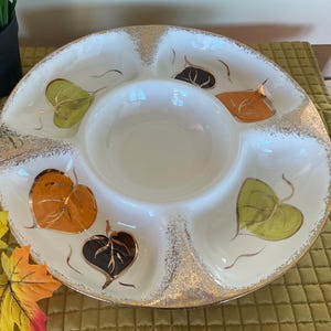 Könnte beinhalten: Eine weiße Keramik-Servierschale mit einer zentralen Schüssel und fünf umliegenden Fächern. Die Schale ist mit goldenen Verzierungen und farbenfrohen Blattmotiven in Orange, Grün und Braun verziert. Ein dekoratives Servierstück zum Thema Herbst.