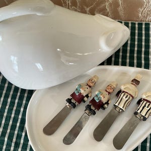Peut inclure: Un plat de service en céramique blanche avec quatre couteaux à beurre. Les couteaux ont des figurines de chef sur les manches. Une grande cruche blanche est en arrière-plan. Les articles sont sur un tissu à carreaux vert et blanc.