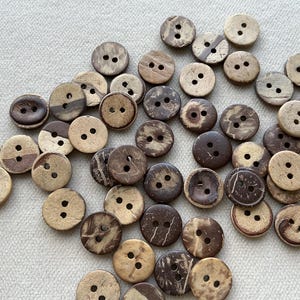 Peut inclure: Un assortiment de boutons ronds de tons naturels, chacun percé de deux trous. Les boutons varient du beige clair au marron foncé, avec des motifs de grain de bois visibles. Idéal pour la couture et les travaux manuels.
