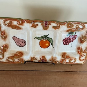 Peut inclure: Plat de service rectangulaire en céramique à trois compartiments. Le plat est blanc cassé avec une bordure décorative dans les tons orange et vert. Chaque section présente un fruit peint : une figue, une pomme et des raisins.