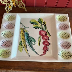 Puede incluir: Una fuente rectangular de cerámica blanca con un diseño de verduras pintado a mano. La fuente presenta una ilustración central de pimientos amarillos y verdes y rábanos rojos. Los bordes tienen hendiduras en forma de hoja, pintadas en tonos verdes, rosas y amarillos.