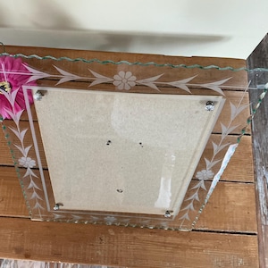 Op de afbeelding: Transparante glazen fotolijst met geëtste bloemen- en bladerontwerpen. De lijst heeft een unieke vorm met een geschulpte rand en wordt bij elkaar gehouden met zilverkleurige hardware. Een roze bloem is naast de lijst geplaatst.