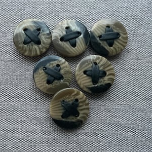 Peut inclure: Six boutons ronds avec un motif camouflage dans les tons de vert, marron et noir. Chaque bouton a un détail de point de croix noir au centre. Les boutons sont disposés sur un fond de tissu gris.