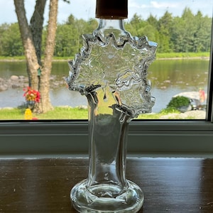 Peut inclure: Une bouteille en verre transparent en forme de feuille d'érable avec une base cylindrique et un petit bouchon à vis marron. Le verre présente un motif de feuille d'érable texturé et en relief sur la partie supérieure, se rétrécissant vers le col.