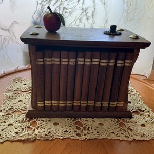 May include: A rectangular wooden box designed to resemble a row of antique books with dark brown spines and gold-coloured horizontal bands. The top surface features four brass-coloured corner accents and a small red apple-shaped decorative object.