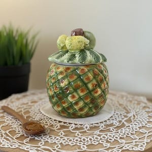 May include: A ceramic pineapple-shaped jar with a lid, featuring green and orange detailing. The lid is adorned with fruit-shaped embellishments. The jar sits on a white doily atop a wooden surface, with a small wooden spoon nearby. A potted plant is in the background.