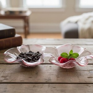 Könnte beinhalten: Zwei rosa, blütenförmige Glasschalen auf einem Holztisch. Eine Schale enthält dunkle, schokoladenüberzogene Espressobohnen, die andere frische Himbeeren und Minze. Die Schalen befinden sich vor einem verschwommenen Hintergrund.