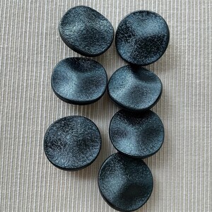 Peut inclure: Sept objets décoratifs ronds noirs et texturés sont disposés sur une surface beige clair à rayures verticales. Les objets ont une forme légèrement irrégulière et un aspect moucheté, suggérant une origine naturelle ou artisanale.