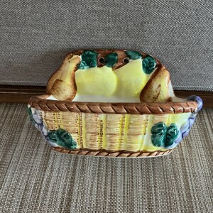 Peut inclure: Plat en céramique en forme de panier avec un motif de poires et de fruits. Le panier a une texture tissée avec des tons marron et jaune, et est décoré de feuilles vertes et de raisins violets. Les poires sont jaunes et marron.
