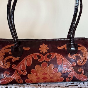 Peut inclure: Un sac à main rectangulaire marron foncé orné d'un motif floral et de volutes en relief de couleur beige clair sur le devant. Le sac comprend deux anses assorties marron foncé à la finition texturée et une fermeture éclair sur le dessus.
