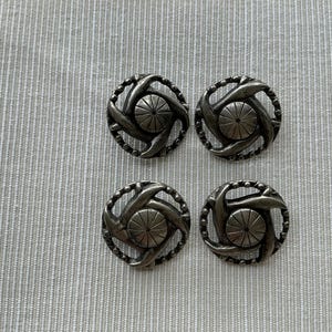 Peut inclure: Quatre boutons argentés antiques. Chaque bouton présente un motif circulaire central texturé entouré d'un motif tourbillonnant en forme de corde. Les boutons sont disposés sur un tissu gris clair texturé.