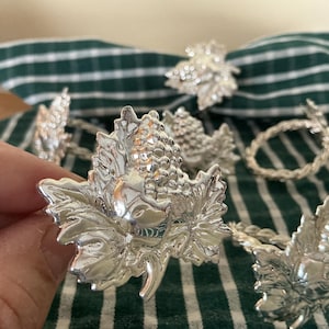 May include: Silver napkin rings featuring a leaf and grape motif. The rings are displayed on a green and white striped cloth. The intricate leaf and grape design adds a touch of sophistication to any dining table. Each ring is a decorative element.