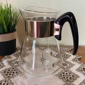 May include: A clear glass coffee carafe featuring a black starburst pattern, a silver-toned metal band around the neck, and a black plastic handle. The glass body includes printed text instructions regarding use on an electric range.