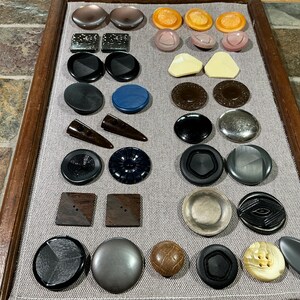 May include: A selection of buttons in diverse shapes and hues. The assortment features round, square, and oval buttons in shades of black, brown, blue, beige, and grey. Some buttons display textured surfaces or distinctive patterns, offering a range of styles.