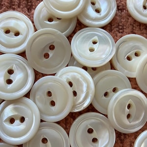 Peut inclure: Un ensemble de boutons ronds, blanc cassé, chacun percé de deux trous. Les boutons présentent un aspect nacré et sont disposés sur une surface texturée, brun rougeâtre. Parfaits pour la couture et les travaux manuels.