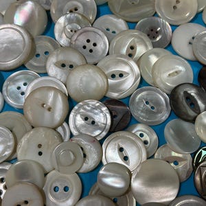 May include: A selection of white and cream-coloured buttons, many with a pearlescent finish. The buttons have two or four holes and come in various sizes and shapes. Some buttons are dark brown or black.