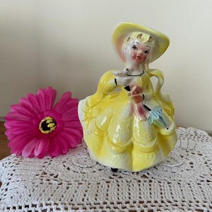 Peut inclure: Figurine en céramique vintage d'une femme en robe et chapeau jaunes, tenant un parasol. La figurine est posée sur un napperon en dentelle blanche, avec une fleur rose vif à proximité. Esthétique classique et antique.