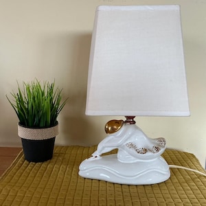 Peut inclure: Lampe de table en céramique blanche avec abat-jour crème. La base de la lampe présente une figurine dorée. Un petit pot noir avec de l'herbe verte artificielle est à gauche de la lampe.