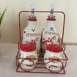 Puede incluir: Un juego de aceite y vinagre de cerámica blanca con detalles rojos y la palabra "Provence". El juego incluye dos botellas con boquillas y dos saleros y pimenteros, todo en un soporte de metal rojo.