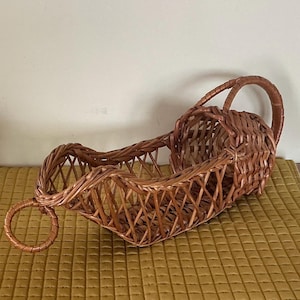 May include: A brown wicker wine bottle holder featuring a woven lattice design, a circular handle at the front, and a curved cradle structure. The basket is constructed from thin, flexible reeds with a glossy finish, designed to hold a bottle.