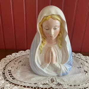 Peut inclure: Buste en céramique de la Vierge Marie, avec un voile blanc et une robe bleue. La figure a les yeux fermés et les mains jointes en prière. Les cheveux sont blonds et le visage est peint avec des traits délicats. Le buste est sur un napperon en dentelle blanche.