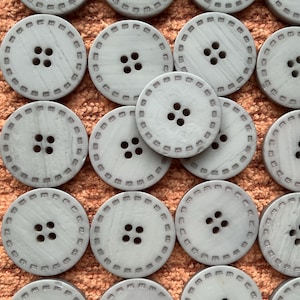 May include: A collection of light gray, round buttons with four holes and a decorative stitched border. The buttons are arranged on a textured, orange-brown surface, showcasing their uniform design and potential for sewing or craft projects.