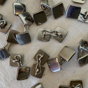 May include: A collection of silver-toned cufflinks with a square design. The cufflinks are scattered across a light-colored surface, showcasing their reflective, polished finish. The cufflinks are a classic accessory.