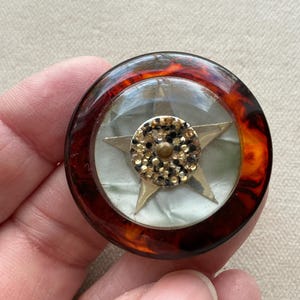 May include: A circular vintage brooch with a tortoiseshell-effect border. The centre has a gold star, a circle of sparkling embellishments, and a pearlescent background. A unique and eye-catching accessory.