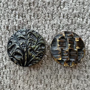 Puede incluir: Dos botones redondos decorativos. Un botón presenta un diseño floral en negro y dorado. El otro botón tiene un patrón geométrico en negro y dorado. Ambos botones están sobre una superficie gris texturizada.