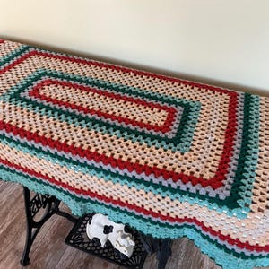 Peut inclure: Une nappe rectangulaire au crochet faite à la main. La nappe présente un motif répétitif de carrés crème, rouges, turquoise et gris. Les bords sont festonnés avec une bordure turquoise. La nappe est exposée sur une table noire.