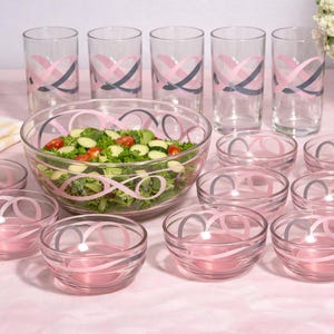 Peut inclure: Un service de table en verre transparent avec un motif de ruban rose et gris. L'ensemble comprend un grand saladier, de petits bols et de grands verres. Le saladier est rempli d'une salade fraîche. L'ensemble est disposé sur une surface rose.