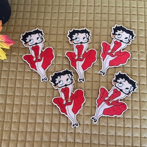 Peut inclure: Cinq écussons Betty Boop. Chaque écusson représente le personnage de dessin animé dans une robe rouge avec un contour noir. Les écussons sont sur une surface texturée brun doré. Les écussons sont dans diverses poses.