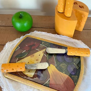Peut inclure: Un plateau à fromage décoratif avec deux couteaux à fromage, une pomme verte et des décorations en forme de fromage. Le plateau présente un motif de fruits. Les couteaux ont des manches orange.