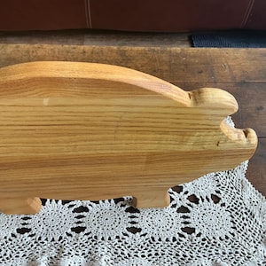 May include: A wooden serving board in the shape of a pig. The board is crafted from light-coloured wood, displaying the grain. The pig has a rounded back, a snout, and small legs. It rests on a white lace doily.