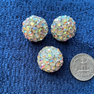 Peut inclure: Trois boucles d'oreilles à clip en strass irisés. Les boucles d'oreilles sont rondes et recouvertes de strass colorés. Une pièce de monnaie américaine de 25 cents est à côté des boucles d'oreilles pour la comparaison de taille.