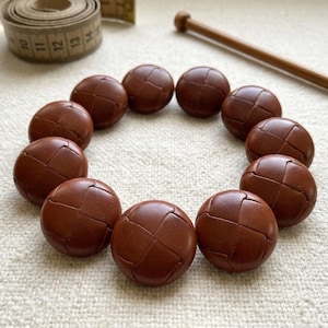 Peut inclure: Onze boutons ronds en cuir marron, avec un motif tressé, disposés en cercle. Une aiguille à tricoter en bois et un mètre ruban sont également présents. Idéal pour la couture.