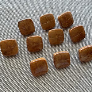 Puede incluir: Diez botones cuadrados de color ámbar con un patrón texturizado en forma de rejilla. Los botones están dispuestos sobre una superficie gris texturizada. Estos botones podrían usarse para coser o hacer manualidades.
