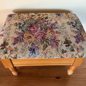 May include: A rectangular footstool featuring a beige fabric top with a woven floral pattern in shades of pink, purple, and green. The base is constructed from light-toned wood and is supported by four turned legs with a rounded, bulbous design.
