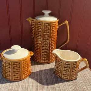 Peut inclure: Un service à café en céramique de trois pièces avec un motif de tissage en panier. L'ensemble comprend une cafetière avec un couvercle, une sucrière avec un couvercle et une crémière. Les pièces sont marron avec des accents blancs.