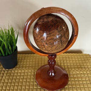 Peut inclure: Un globe terrestre en bois avec une finition marron foncé, monté sur un support assorti. Le globe est entouré d'un anneau en bois. Le globe présente des détails géographiques et des noms de lieux dessinés à la main. Le support est rond et possède une tige décorative.