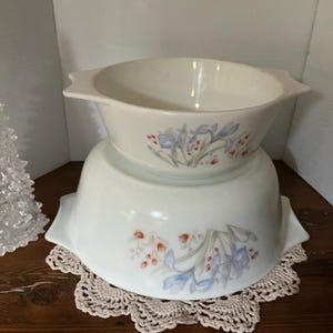 May include: Two off-white ceramic bowls with handles, stacked on a crocheted doily. The bowls have a floral design with blue, red, and green accents. A clear, decorative tree is on the left.