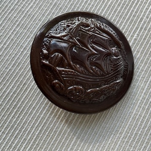 Puede incluir: Un botón redondo marrón oscuro con un relieve detallado de un velero. El botón está sobre un fondo de tela texturizada gris claro. El diseño del barco incluye velas, olas y detalles intrincados.