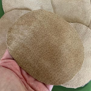 May include: Close-up of several tan suede leather circles. One is held in a hand, showing the texture and color variations. The circles are of varying sizes and shapes, with a soft, matte finish. The background is a green surface.