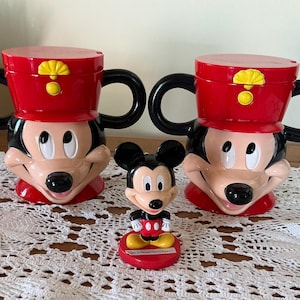 May include: Two red and black plastic mugs shaped like Mickey Mouse wearing a soldier hat with yellow accents, accompanied by a small Mickey Mouse figurine on a red base with the text Walt Disney World Resort printed on the front of the base.