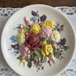 May include: A white ceramic plate featuring a raised floral design. The flowers are in shades of yellow, pink, and burgundy, with green leaves and accents of blue and purple. The plate has a scalloped edge.