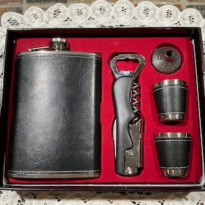 May include: A black leather-wrapped hip flask and bar tool set. The set includes a flask, two shot glasses, a bottle opener, and a funnel, presented in a black box with a red interior. The shot glasses and flask have silver accents.