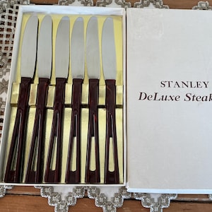 Peut inclure: Un coffret de six couteaux à steak. Les couteaux ont des lames argentées et des manches bordeaux foncés. La boîte blanche porte l'inscription "STANLEY Deluxe Steak Set". Les couteaux sont disposés dans un insert doré.