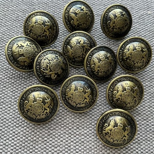 Peut inclure: Ensemble de boutons antiques de couleur laiton. Chaque bouton présente un motif en relief détaillé avec un emblème central et des éléments décoratifs. Les boutons ronds ont un aspect légèrement vieilli.